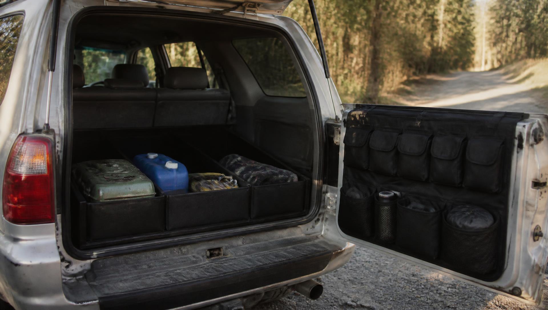 Open rear cargo area of a 4Runner on a gravel road: black cargo organiser holding a jerry can, water jug and blanket, with a hanging tailgate organiser on the open hatch